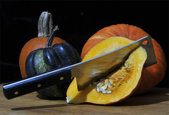 No mercy for pumpkins An assortment of pumpkins in different colours. One has been halved and a cleaver is stuck deeply into its flesh.