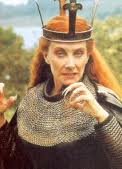 a pale skinned woman with long red hair is wearing a crown and a chainmail shirt, holding up one hand with very long fingernails and looking angry