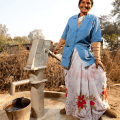 A middle aged woman with dark hair wearing a sari and a jacket over the top poses by a well pump that she maintains