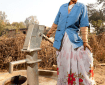 A middle aged woman with dark hair wearing a sari and a jacket over the top poses by a well pump that she maintains