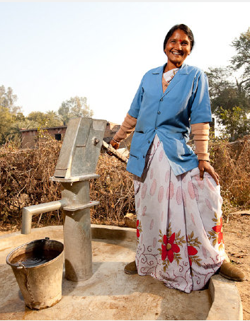 A middle aged woman with dark hair wearing a sari and a jacket over the top poses by a well pump that she maintains
