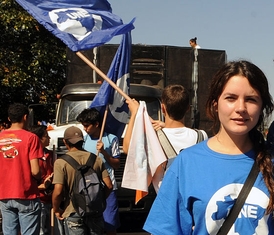 A young woman with pale skin and dark hair is in the left foreground, wearing a blue T-shirt with a white logo, behind her are many other students waving flags and marching