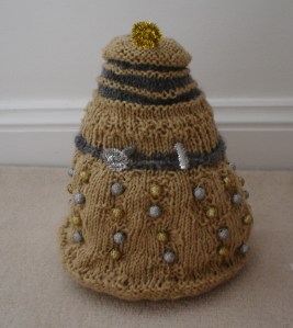 A gold Dalek with silver accents and gold and silver beads as its bobbles. The plunger and ray gun are made of silver pipecleaners, and the eyestalk out of a gold one.
