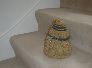 A gold Dalek with silver accents and gold and silver beads as its bobbles. The plunger and ray gun are made of a silver pipecleaner, and the eyestalk out of a gold one. The Dalek is posed on some stairs.