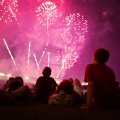 In the foreground we see the silhouettes of seated people, watching the fireworks show in the distance