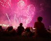 In the foreground we see the silhouettes of seated people, watching the fireworks show in the distance