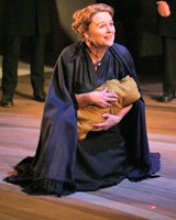 A middle-aged white woman in a black cloak kneels, holding a baby wrapped in brown cloth.