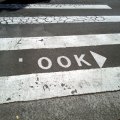 Photo of a pedestrian crossing, where there is normally writing that says LOOK with an arrow pointing to the right. The L has been rubbed off so that the sign now says OOK.