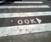 Photo of a pedestrian crossing, where there is normally writing that says LOOK with an arrow pointing to the right. The L has been rubbed off so that the sign now says OOK.