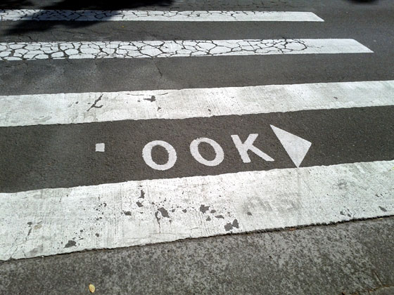 Photo of a pedestrian crossing, where there is normally writing that says LOOK with an arrow pointing to the right. The L has been rubbed off so that the sign now says OOK.