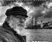 a silver bearded man in a fishing cap gazes thoughtfully into the distance, in the background is a wide stretch of water and a square-rigged sailing ship beside a lighthouse, all underneath a dark and cloudy sky
