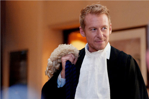 A pale skinned man with blond hair is wearing a barrister's silks and holding his court wig over one shoulder while looking sideways out of frame
