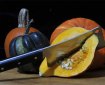An assortment of pumpkins in different colours. One has been halved and a cleaver is stuck deeply into its flesh.