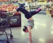 A woman in practical yoga clothes is doing a handstand in the middle of the fruit/veg aisle at market - one leg rests backwards on the shopping trolley while the other leg is fully extended elegantly forwards