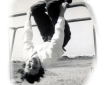 black and white photo of a young woman with pale skin and short dark curly hair. She is hanging upside down by her knees from playground "jungle bars", laughing at the camera.