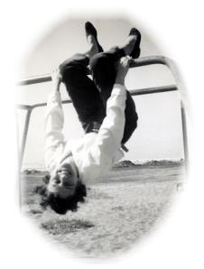 black and white photo of a young woman with pale skin and short dark curly hair. She is hanging upside down by her knees from playground "jungle bars", laughing at the camera.