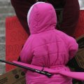 A very small child in a bright pink parka, viewed from behind, with a bright pink rifle slung on her back.