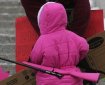 A very small child in a bright pink parka, viewed from behind, with a bright pink rifle slung on her back.