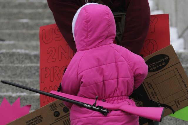 A very small child in a bright pink parka, viewed from behind, with a bright pink rifle slung on her back.