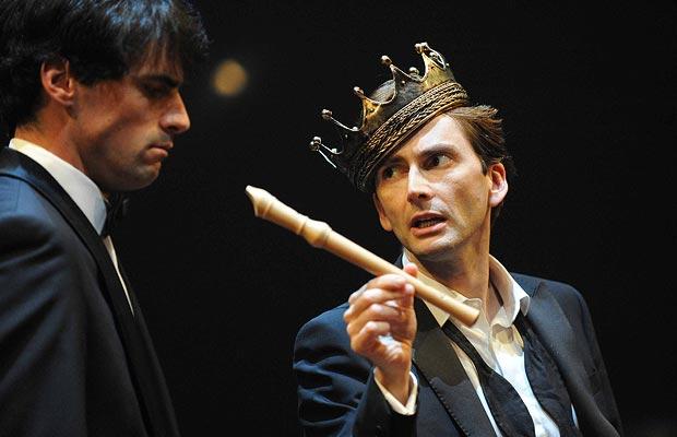 David Tennant in a suit, tie undone, with a crown tipped over one eye, offers his fellow player a recorder.