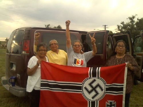 Four elderly but strong looking indigenous American women stand next to a van. They are holding a captured Nazi flag and cheering in preparation for burning it.