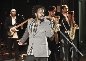 Black man at microphone with 3 men behind (2 tenor sax, 1 electric guitar).