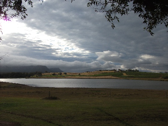A dam in the foreground, a vine covered low hill, then a mountain range to the left and a dramatic cloudy sky with a few blue patches to the right