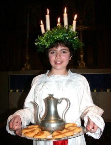 Girl of about 12 wearing crown of leaves and candles, carrying tray with pewter jug and buns.