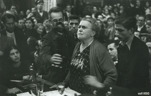 B&W of elderly woman standing at a dest speaking into a microphone, surrounded by people.