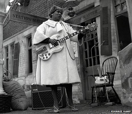 B&W of large black woman in fluffy 1960s coat playing a white electric guitar.