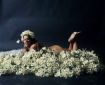 A paleskinned fat woman with dark hair lies front-down and naked upon a bed of white daisy-like flowers. Her head is held high with a wide smile looiing into the camera. We can see her head, one shoulder, a bit of buttock, and her feet up in the air. One hand is just visible holding a flower next to her face, and we see another flower held between her toes so it lies just on the sole of one foot.
