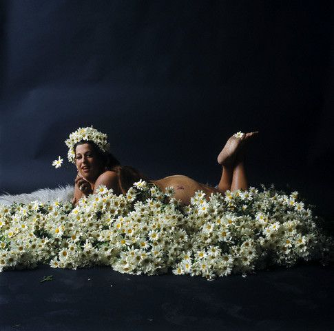 A paleskinned fat woman with dark hair lies front-down and naked upon a bed of white daisy-like flowers. Her head is held high with a wide smile looiing into the camera. We can see her head, one shoulder, a bit of buttock, and her feet up in the air. One hand is just visible holding a flower next to her face, and we see another flower held between her toes so it lies just on the sole of one foot.