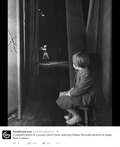 B&W photo of a toddler girl on a stool watching a woman on stage in the distance.