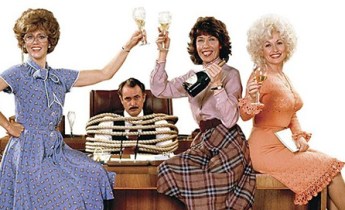 Three woman sit on a large business desk, toasting each other with champagne. Seated behind them is their male boss, tied to his office chair.